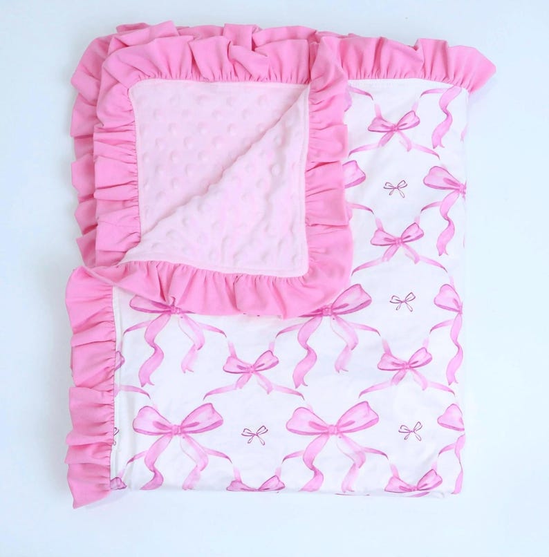 Puede incluir: Una manta rosa para beb&eacute; con fondo blanco y estampado de lazos rosas. La manta tiene un borde rosa con volantes y un interior rosa suave y texturizado. Dise&ntilde;ada para beb&eacute;s y ni&ntilde;os peque&ntilde;os.