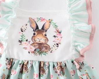 May include: A white sleeveless dress with a light blue and white striped ruffle trim on the straps. The dress has a floral wreath with a brown bunny in the centre. The bottom of the dress is light blue with a floral and bunny pattern.