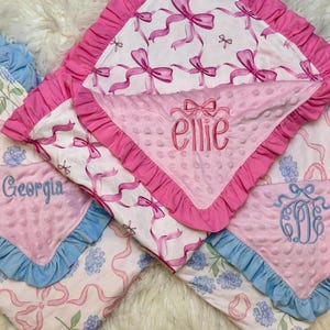 May include: Several baby blankets with pink and blue accents. The blankets feature floral and ribbon patterns, with some personalized with names like "Ellie" and "Georgia". Each blanket has a ruffled edge.