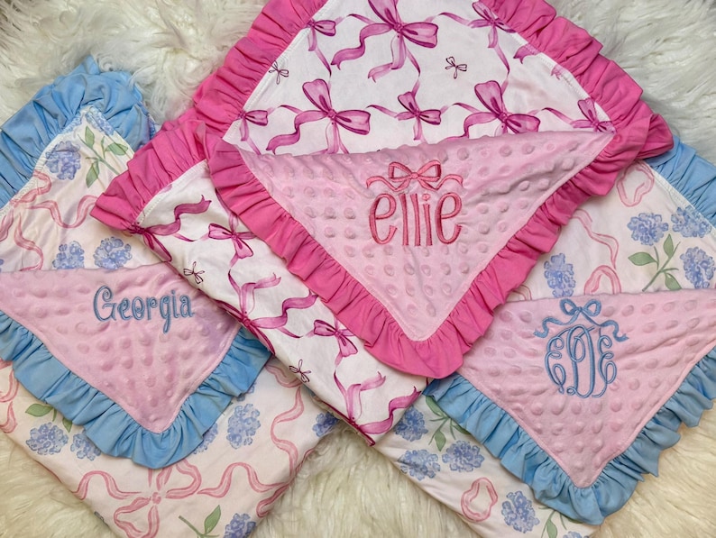 Puede incluir: Varias mantas para beb&eacute;s con detalles en rosa y azul. Las mantas presentan dise&ntilde;os florales y de lazos, algunas personalizadas con nombres como "Ellie" y "Georgia". Cada manta tiene un borde con volantes.