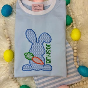 K&ouml;nnte beinhalten: Hellblaues Langarmshirt und gestreifte Hose. Das Shirt zeigt ein blaues Hasenmuster mit einer Karotte und dem Namen "Joshua" in Gr&uuml;n. Das Shirt hat einen wei&szlig;en Kragen und den Text "Rags to Riches Monogram".