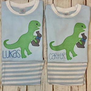 May include: Two light blue t-shirts with white stripes, featuring a green dinosaur holding a chocolate bunny. The shirts are personalized with the names "Lukas" and "Carter".