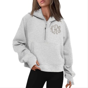 May include: A light gray hooded sweatshirt with a half-zip closure and a kangaroo pocket. The sweatshirt features a monogram in a lighter shade of gray on the upper chest. The sleeves have thumb holes.