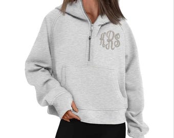 May include: A light gray hooded sweatshirt with a half-zip closure and a kangaroo pocket. The sweatshirt features a monogram in a lighter shade of gray on the upper chest. The sleeves have thumb holes.