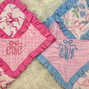 Puede incluir: Dos mantas para beb&eacute;. Una es rosa con un lazo rosa y el nombre "ellie" bordado en rosa. La otra es rosa con ribete azul y las iniciales "EWH" bordadas en azul. Ambas tienen un patr&oacute;n de lazo blanco.