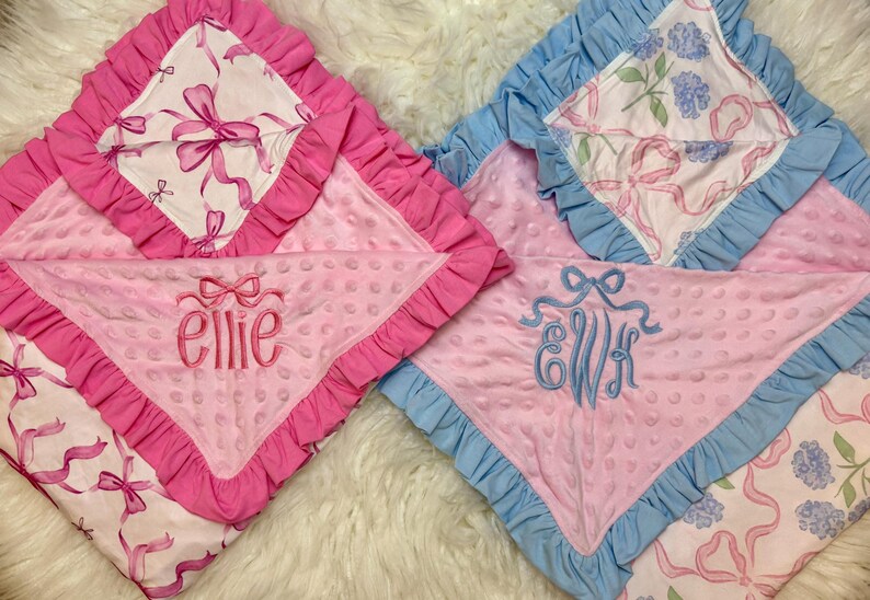 Puede incluir: Dos mantas para beb&eacute;. Una es rosa con un lazo rosa y el nombre "ellie" bordado en rosa. La otra es rosa con ribete azul y las iniciales "EWH" bordadas en azul. Ambas tienen un patr&oacute;n de lazo blanco.
