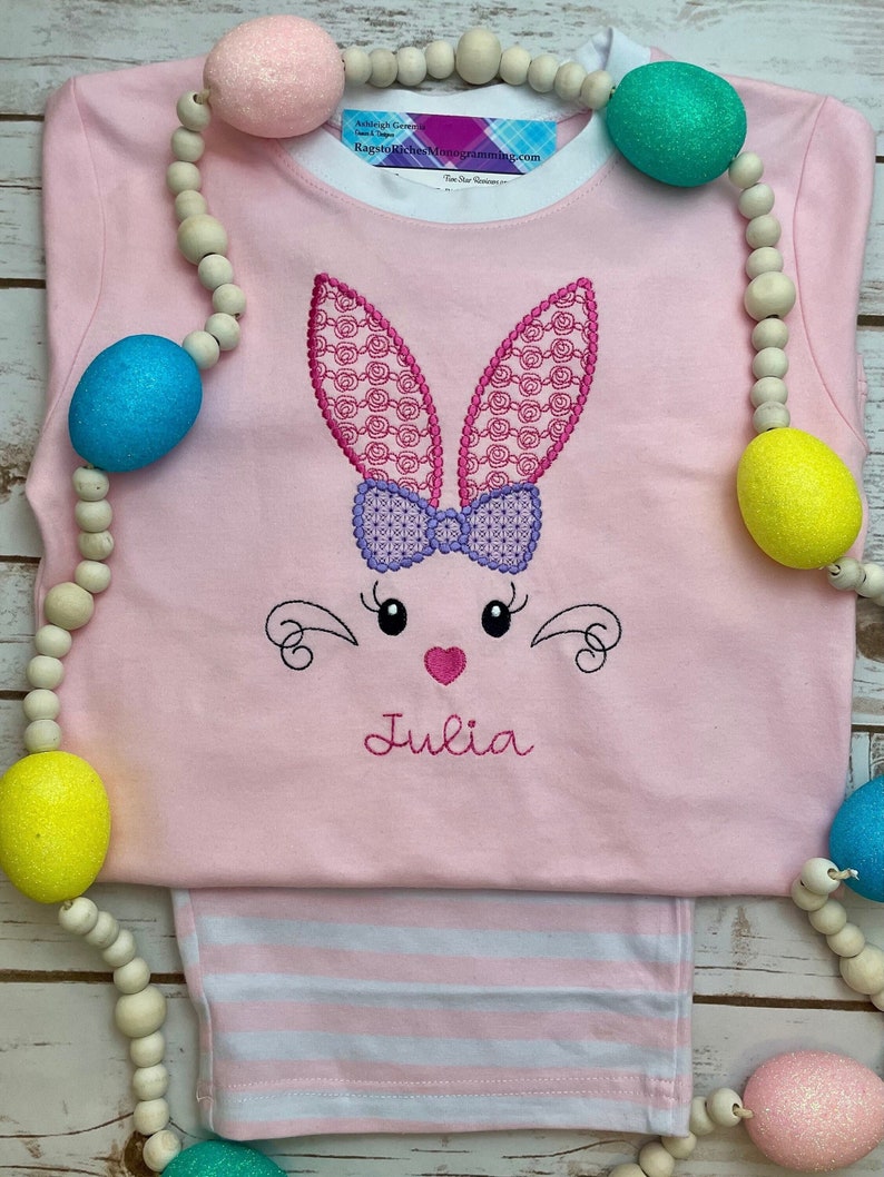 K&ouml;nnte beinhalten: Rosa Langarmshirt und gestreifte Hose mit einem gestickten Hasengesicht-Design. Der Hase hat rosa Ohren mit Muster, eine lila Schleife und den Namen "Julia" darunter gestickt. Eine Holzperlen-Girlande mit bunten Ostereiern umgibt das Set.