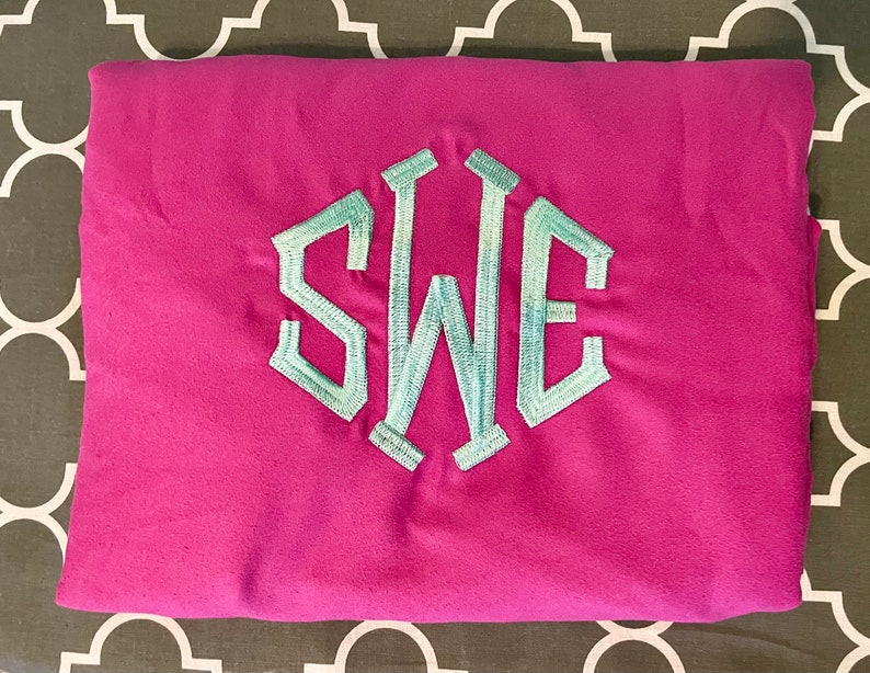 May include: A folded pink t-shirt with a light blue embroidered monogram of the letters "SWE" in a hexagonal shape.