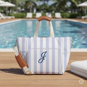 May include: A tote bag featuring a light blue and white vertical striped pattern with a dark blue embroidered letter J on the front pocket. The bag includes cream-colored fabric handles and a detachable shoulder strap with brown leather accents.