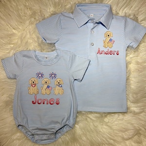 May include: Two light blue striped baby outfits. One is a polo shirt with a puppy holding an American flag and the name "Anders" embroidered in red. The other is a romper with three puppies, fireworks, and the name "Jones" embroidered in red.