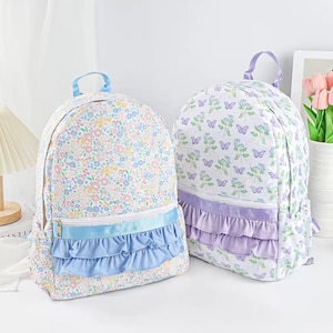 May include: Two floral print backpacks. One backpack has a light blue ruffle and trim, the other has a lavender ruffle and trim. Both have a top handle and a zipper closure. The floral patterns include butterflies and flowers.