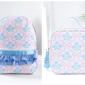 May include: A white backpack and lunch bag set. Both items feature a repeating pattern of pink bows, blue flowers, and green leaves. The backpack has a blue ruffle detail and the lunch bag has a blue shoulder strap.
