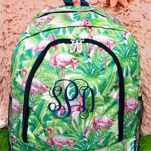 May include: A green and white backpack with a tropical flamingo and leaf pattern. The backpack has a navy blue zipper and handle, and a monogram in navy blue. The backpack has a mesh side pocket.