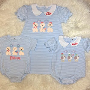 May include: Three light blue striped baby outfits with white collars. One is a dress with the name "Ellie" embroidered on it, and the other two are rompers with the names "Beau" and "Amelia". Each outfit features embroidered patriotic designs.