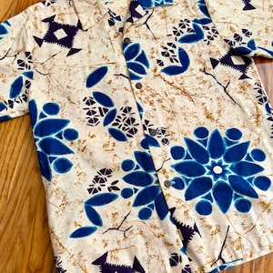 May include: A short-sleeved button-down shirt featuring a cream-colored base with a pattern of dark blue geometric shapes, floral-like motifs, and thin brown branch-like lines. The shirt includes a standard collar and dark brown buttons.
