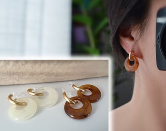 Puede incluir: Pendientes con aro dorado y colgante circular translúcido. Un par es de color crema claro y el otro marrón cálido. Los pendientes se muestran sobre una superficie blanca y se llevan puestos.