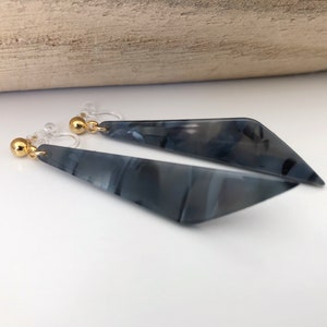 Peut inclure: Une paire de boucles d'oreilles pendantes au design géométrique. Les boucles d'oreilles sont de couleur gris foncé avec un effet marbré. Elles ont des clous dorés et des crochets en plastique transparent.