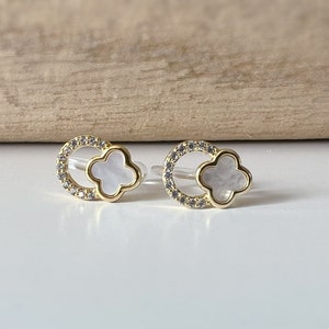 May include: A pair of gold earrings with a white clover-shaped charm surrounded by a circle of clear rhinestones.