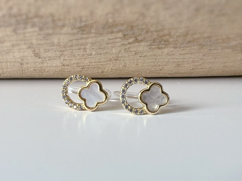 May include: A pair of gold earrings with a white clover-shaped charm surrounded by a circle of clear rhinestones.