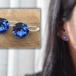Könnte beinhalten: Ein Paar blaue Kristall-Ohrstecker mit durchsichtigen Kunststoff-Rücken. Die Ohrringe sind auf einer Holzoberfläche und am Ohr einer Person zu sehen.