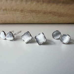 Peut inclure: Plusieurs boucles d'oreilles à clous argentées avec des pierres blanches iridescentes de forme ovale, carrée et ronde. Les boucles d'oreilles sont présentées sur une surface blanche, avec un fond en bois.