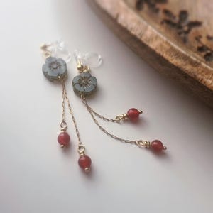 May include: Delicate floral earrings with a light blue flower design and dangling red beads. The earrings have a gold-colored chain and are set against a white background. The earrings are a beautiful accessory.
