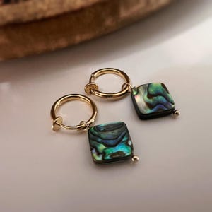 Könnte beinhalten: Ein Paar goldfarbene Creolen mit quadratischen Abalone-Muschel-Anhängern. Die schillernden Anhänger zeigen Farbtöne von Grün, Blau und Schwarz. Die Ohrringe werden auf einer weißen Oberfläche präsentiert.
