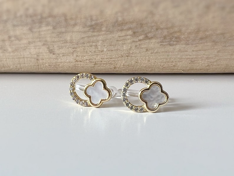 May include: A pair of gold earrings with a white clover-shaped stone set in a circle of clear rhinestones.