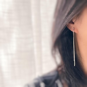 May include: A close-up of a silver earring with a long, slender bar design. The earring is dangling from the ear of a person with dark hair. The background is blurred, with a soft, neutral color palette.