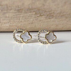 May include: A pair of gold earrings with a white clover-shaped shell in the center of each earring. The earrings are surrounded by a circle of small clear stones.