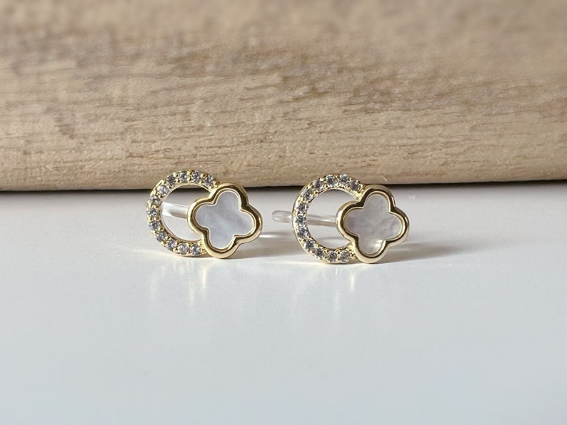 May include: A pair of gold earrings with a white clover-shaped shell in the center of each earring. The earrings are surrounded by a circle of small clear stones.