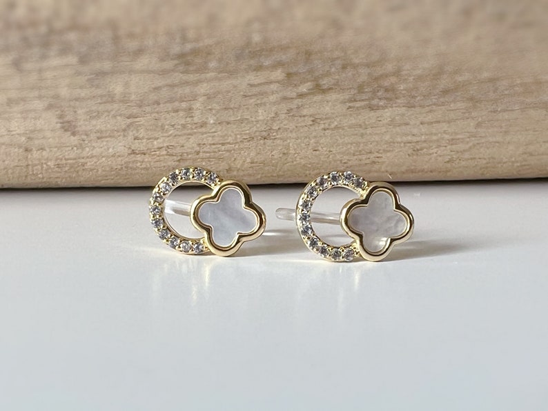 May include: A pair of gold earrings with a white clover design. The clover is surrounded by a circle of small clear stones.