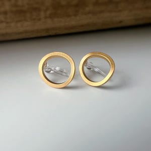 Könnte beinhalten: Ein Paar goldfarbene Ohrringe mit offenem Kreis und transparenten Kunststoff-Steckern. Die Ohrringe sind rund und haben eine matte Oberfläche. Der Hintergrund ist eine hellgraue Oberfläche mit einem Holzelement.