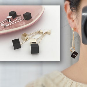 Peut inclure: Une paire de boucles d'oreilles pendantes dorées avec des breloques en forme de cube noir. Les boucles d'oreilles sont présentées sur l'oreille d'un mannequin et sur une surface rose.