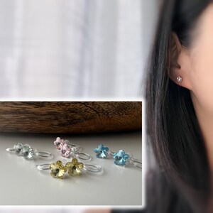以下が含まれることがあります： クリア、ピンク、イエロー、ブルーなど、さまざまな色の花型のクリスタルリングのコレクション。リングは白い表面に表示され、木製の背景があります。女性は花型のイヤリングをしています。