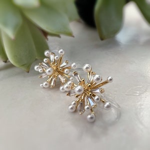 Peut inclure: Une paire de boucles d'oreilles en or avec une explosion de perles blanches et de strass transparents.