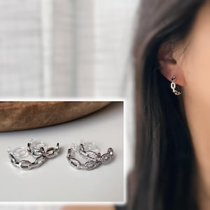 Op de afbeelding: Zilverkleurige oorringen met schakelketting. Eén paar is effen, het andere is bezet met kleine, heldere steentjes. De oorbellen worden getoond op een wit oppervlak en gedragen aan een oor. De oorbellen hebben een moderne, minimalistische stijl.