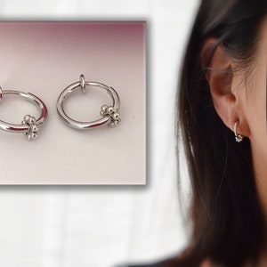 Peut inclure: Une paire de boucles d'oreilles clips argentées avec un petit motif floral. Les boucles d'oreilles sont présentées sur un fond blanc et sur l'oreille d'une personne.