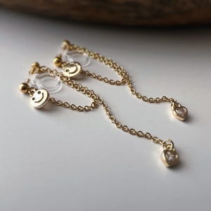 May include: Gold-colored, dangling earrings with a smiley face design and a heart-shaped charm. The earrings have a delicate chain and a clear, plastic backing. The earrings are on a white surface.