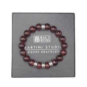 Peut inclure: Un bracelet de perles rondes, rouge foncé, avec des entretoises argentées. Le bracelet est présenté sur une boîte grise foncée avec le texte "BALI BEADS" et "KARTINI STUDIO LUXURY BRACELETS".