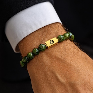 Pode incluir: Pulseira de contas verdes com um elemento retangular dourado. A pulseira é feita de pequenas contas redondas verde-escuras. O elemento dourado tem um símbolo gravado. A pulseira é usada no pulso.