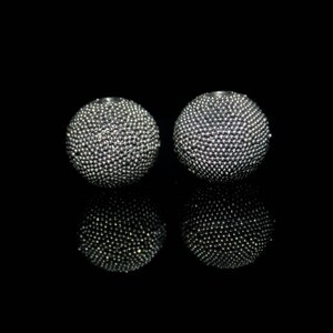 May include: Two silver-colored, spherical earrings with a textured surface, reflecting light. The earrings are set against a black background, creating a contrast that highlights their detailed design. The spheres appear to be made of small, closely packed beads.