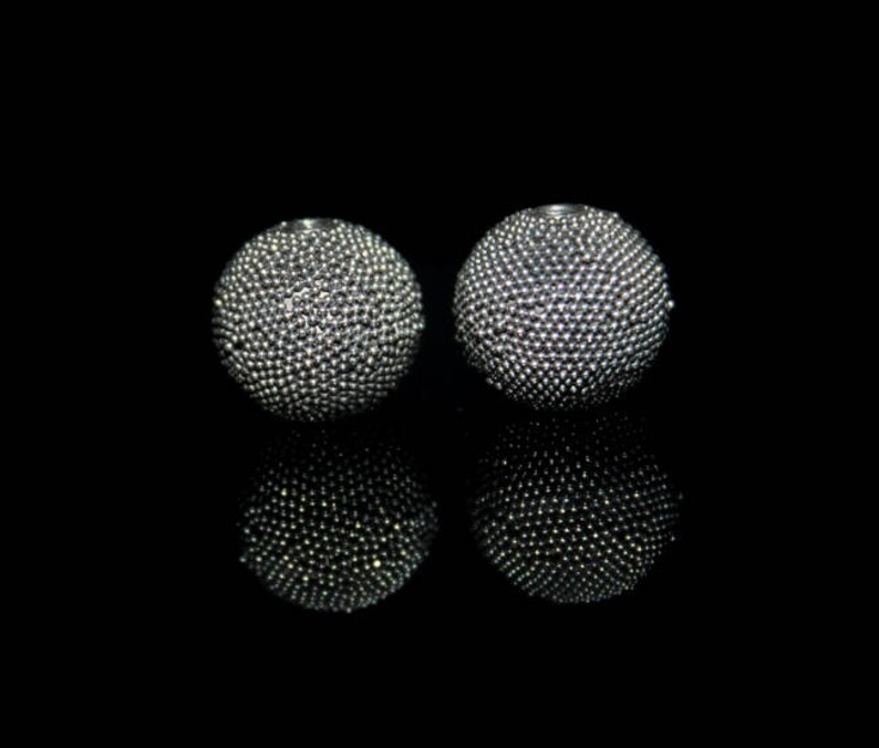 May include: Two silver-colored, spherical earrings with a textured surface, reflecting light. The earrings are set against a black background, creating a contrast that highlights their detailed design. The spheres appear to be made of small, closely packed beads.