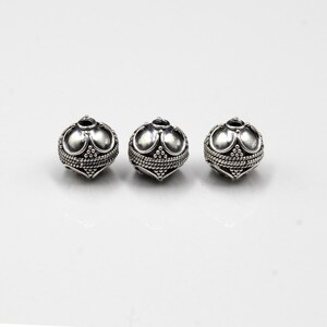 May include: Three silver-colored beads with an ornate design. Each bead has a rounded shape with a textured surface, featuring a pattern of small dots and curved lines. The beads are likely for jewelry making.