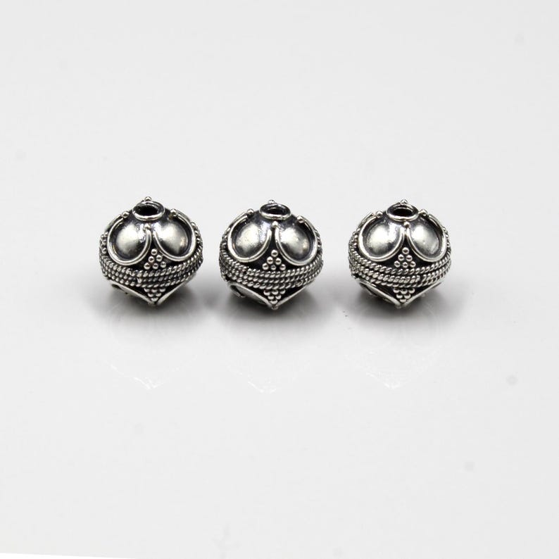 May include: Three silver-colored beads with an ornate design. Each bead has a rounded shape with a textured surface, featuring a pattern of small dots and curved lines. The beads are likely for jewelry making.
