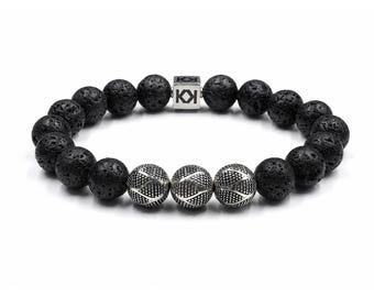 Op de afbeelding: Zwart lava steen kralenarmband met drie zilverkleurige, patroonaccentkralen. De armband heeft een zilverkleurige sluiting met de letters "KI". De kralen zijn rond en hebben een textuur.