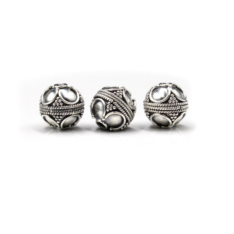 May include: Three silver-colored, spherical beads with intricate designs. Each bead features a pattern of oval shapes and small dots, with a rope-like texture around the center. The beads are displayed against a white background.