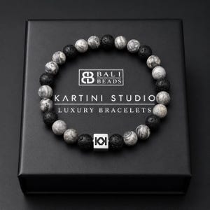 May include: A black box displays a bracelet made of gray and black beads. The bracelet is made of round beads, some with a marbled pattern. The box has the text "BALI BEADS" and "KARTINI STUDIO LUXURY BRACELETS". A silver square with the letters "KI" is also visible.