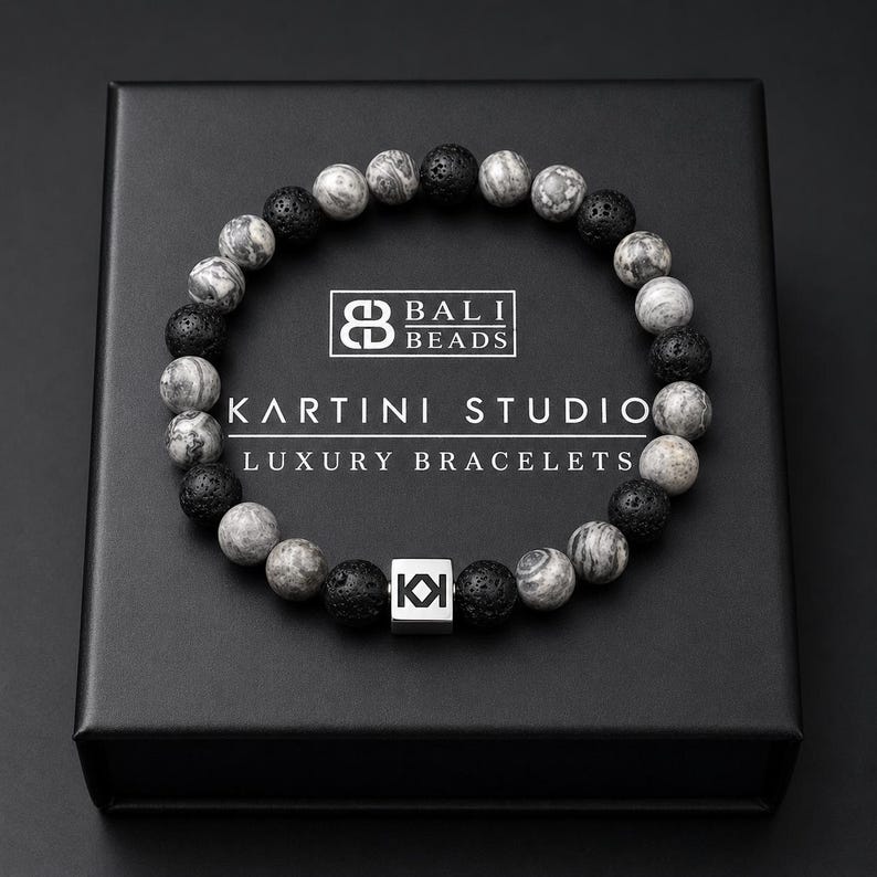 May include: A black box displays a bracelet made of gray and black beads. The bracelet is made of round beads, some with a marbled pattern. The box has the text "BALI BEADS" and "KARTINI STUDIO LUXURY BRACELETS". A silver square with the letters "KI" is also visible.