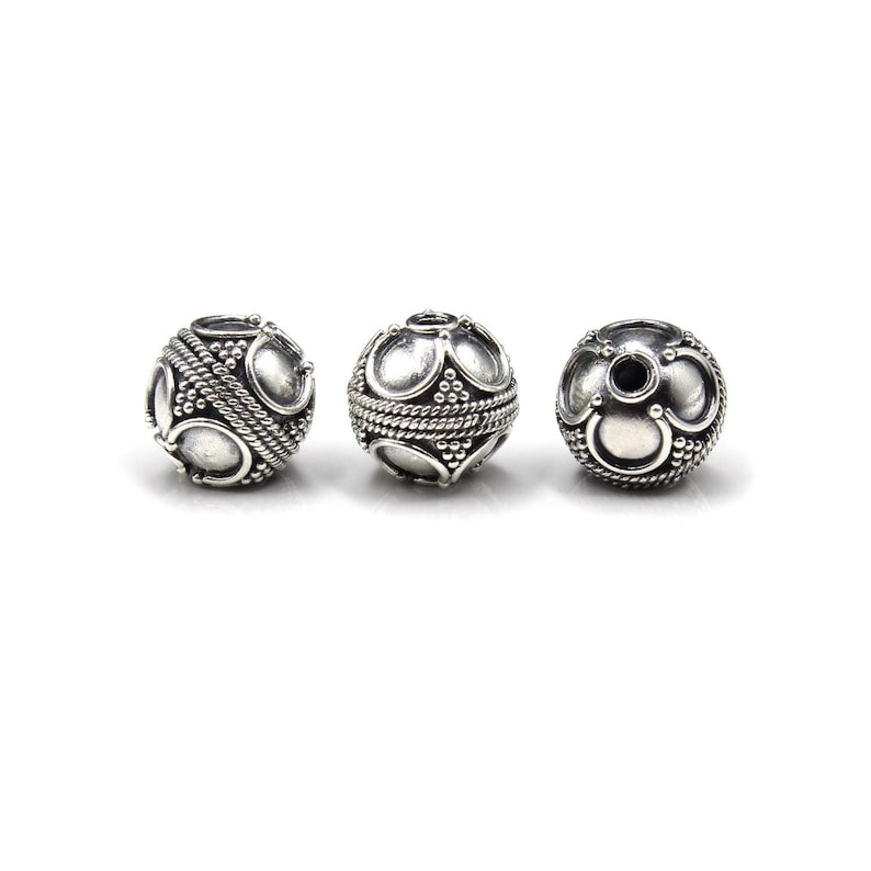 May include: Three silver-colored, round beads with intricate designs. Each bead features a pattern of oval shapes, small dots, and a rope-like texture around the center. The beads are displayed against a white background.